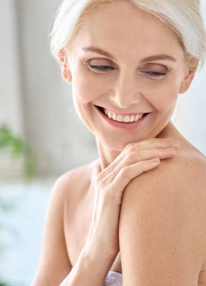 Portrait of happy smiling attractive senior adult nude blonde lady wearing white towel after morning shower touching embracing shoulder with pleasure. Body skin care concept.